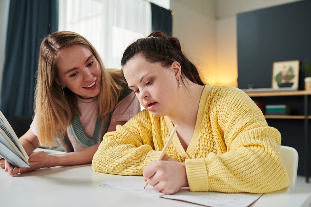 Portrait of young Caucasian woman working as teacher holding textbook giving individual class to girl with Down syndrome